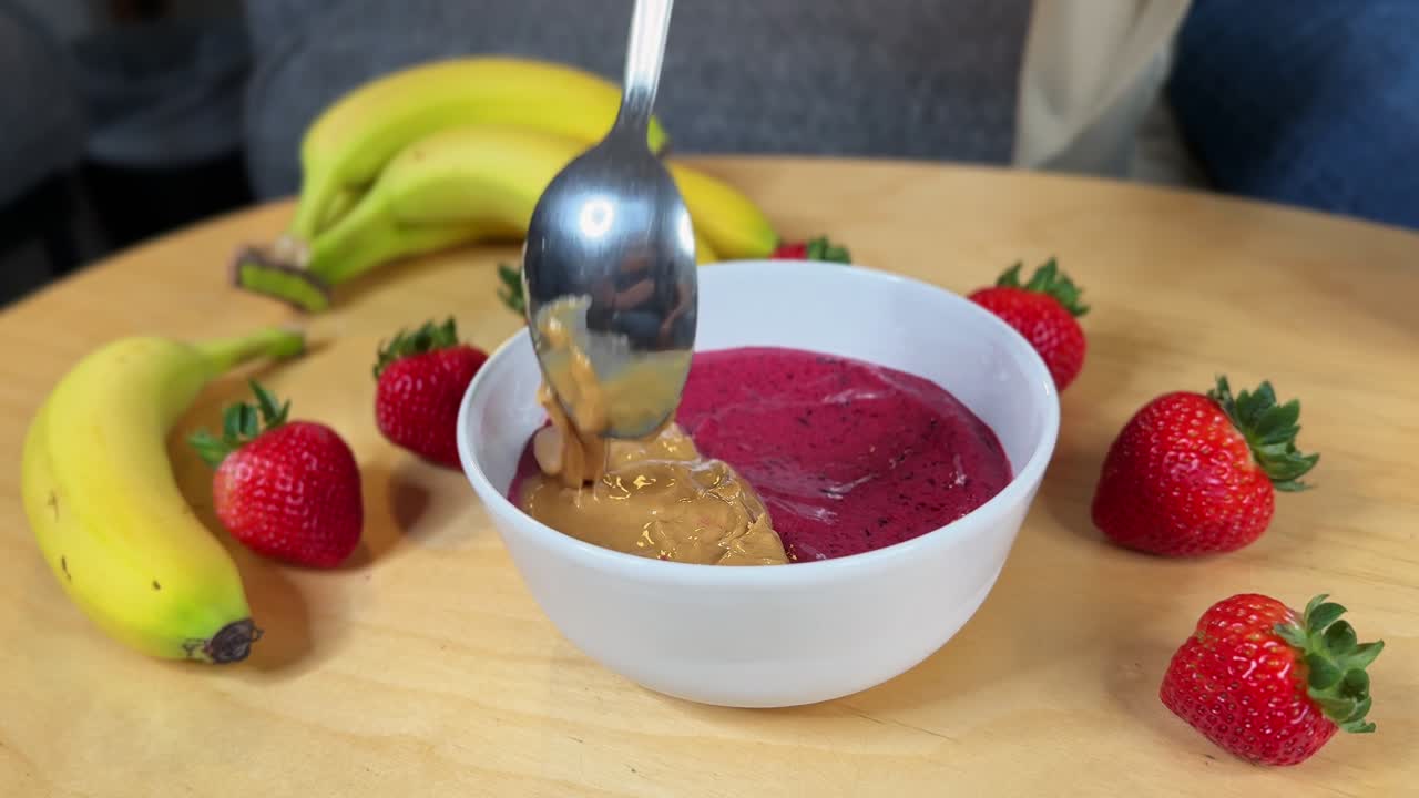 cucchiaio che aggiunge burro di arachidi cremoso in una densa ciotola di frullato di açaí, circondata da fragole e banane fresche: b-roll perfetto per fasi di ricette, colazione sana e promozioni benessere