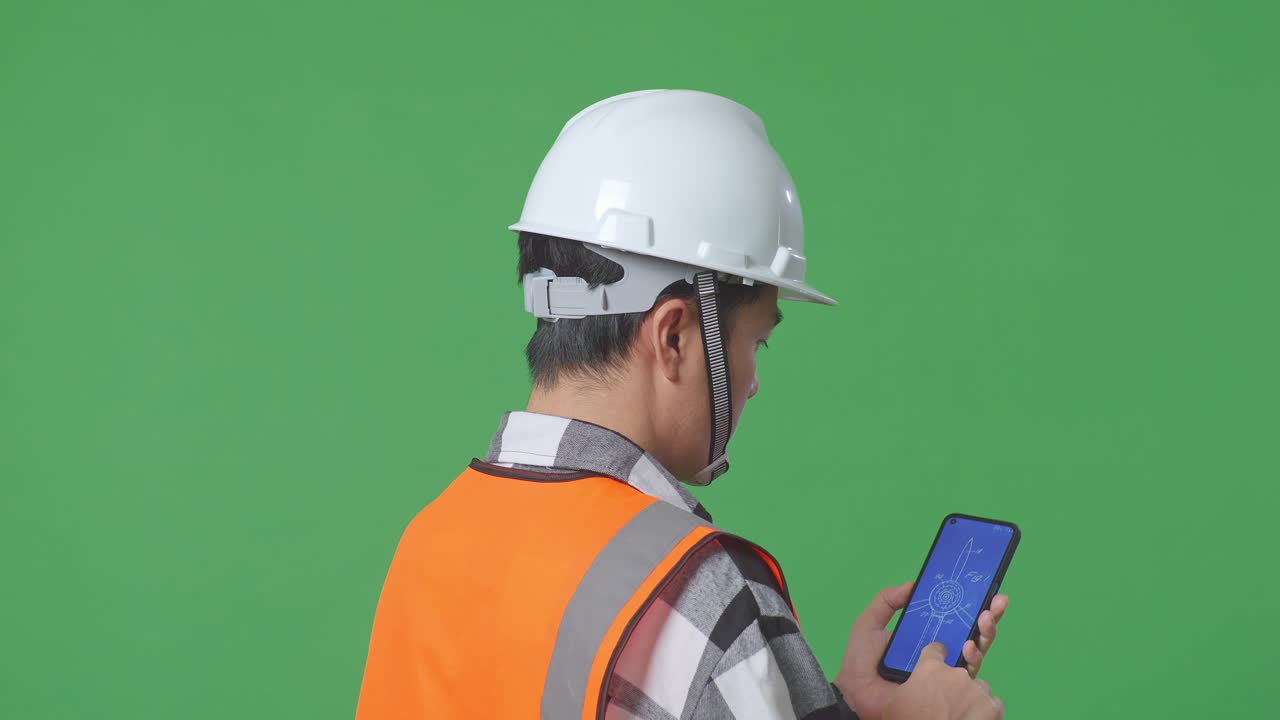 Close Up Back View Of Asian Male Engineer With Safety Helmet Looking At The Wind Turbine Blueprint On Smartphone While Standing In The Green Screen Background Studio