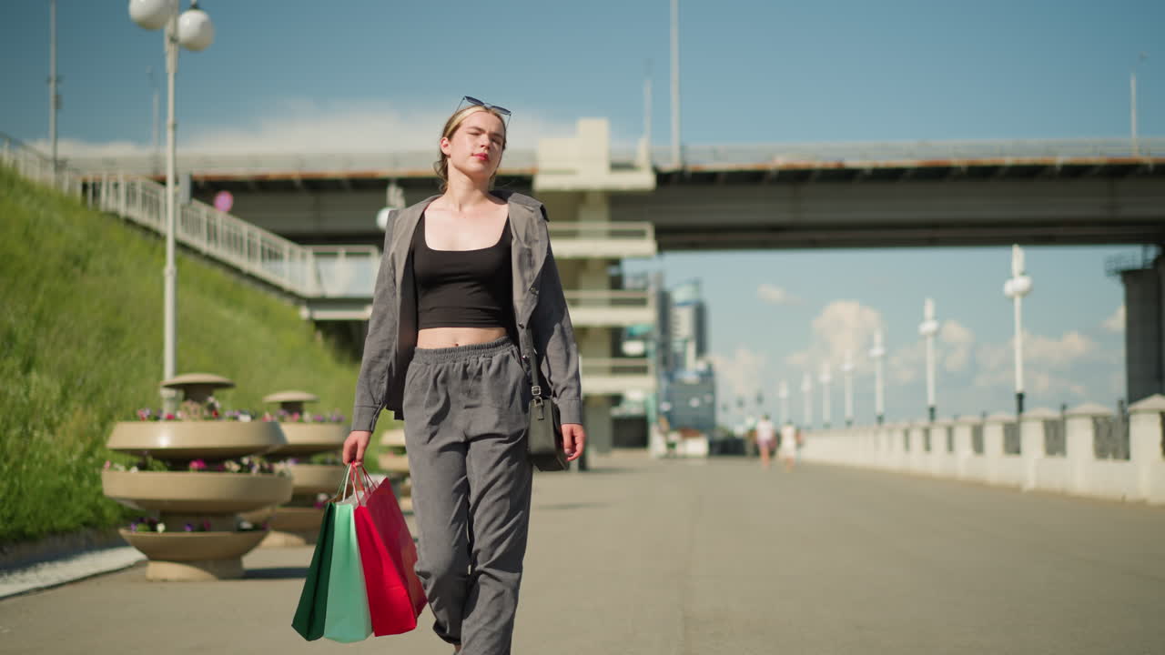 한 여인이 꽃 베스트 근처의 오른손에 쇼핑 가방을 들고 걸어가고, 한 남자가 다리의 레일에 기대어 차가 그녀의 뒤에 있는 오버헤드 다리에서 움직이고 있습니다.