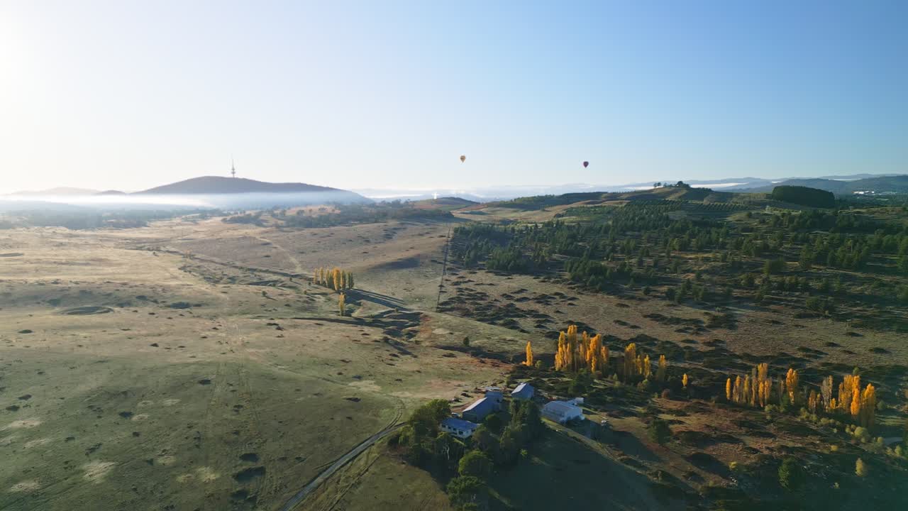 Early morning drone view of two hot air balloons above misty valleys with Canberra’s tower standing tall in the background.
