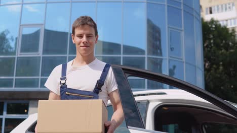 a delivery worker in a white t-shirt and blue jumpsuit stands by an open car and holds a box