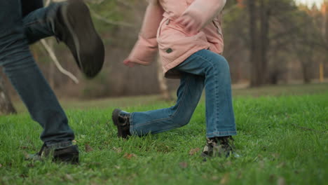 a blurred close-up shot features a girl and a man walking on lush green grass in a park. the girl, wearing a pink jacket, jeans, and black shoes, is in motion, with the background showing trees