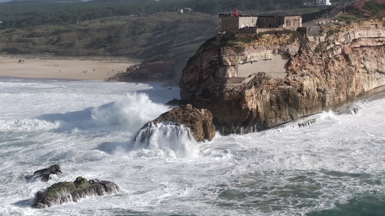 Aerial drone shot of big waves breaking on rocks and cliffs on a day with giant waves in Nazaré, Portugal, Europe. Farol da Nazaré lighthouse and North Beach