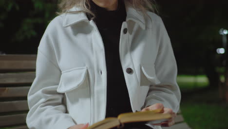 young girl reading alone under streetlight at night as she gently flips through pages of open book, wearing white coat with calm breeze and dark green surroundings