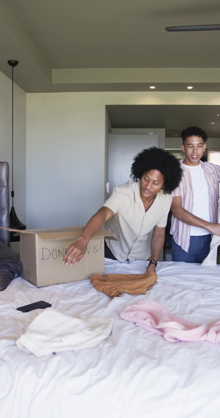 Video verticale: Apertura di una scatola per donazioni, due amici che sistemano vestiti sul letto, controllando il telefono per donare.