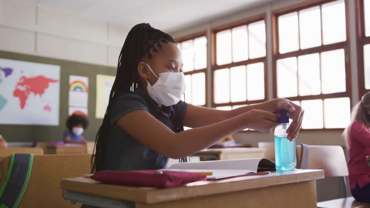 menina usando máscara de rosto desinfetando as mãos enquanto está sentada em sua mesa na escola