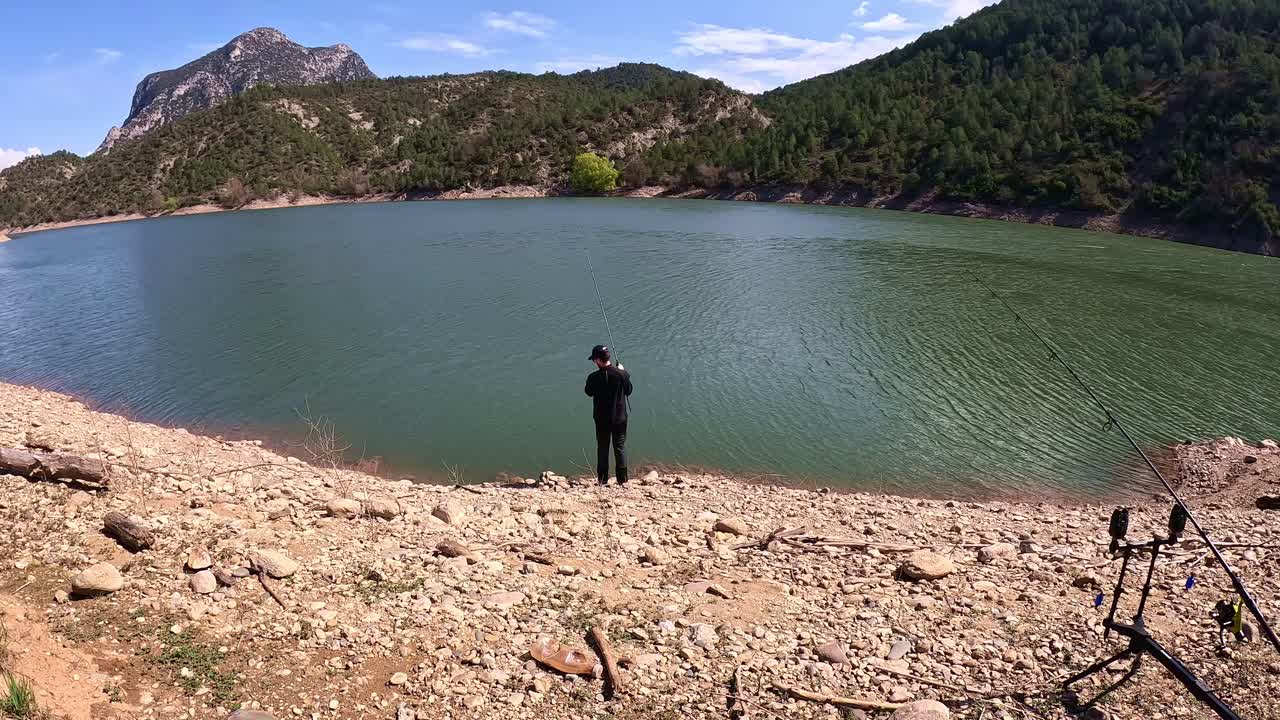 A solitary fisherman stands by a serene mountain lake, focused on capturing a fish. The backdrop is filled with a mix of lush forested hills and a prominent mountain peak under a clear blue sky