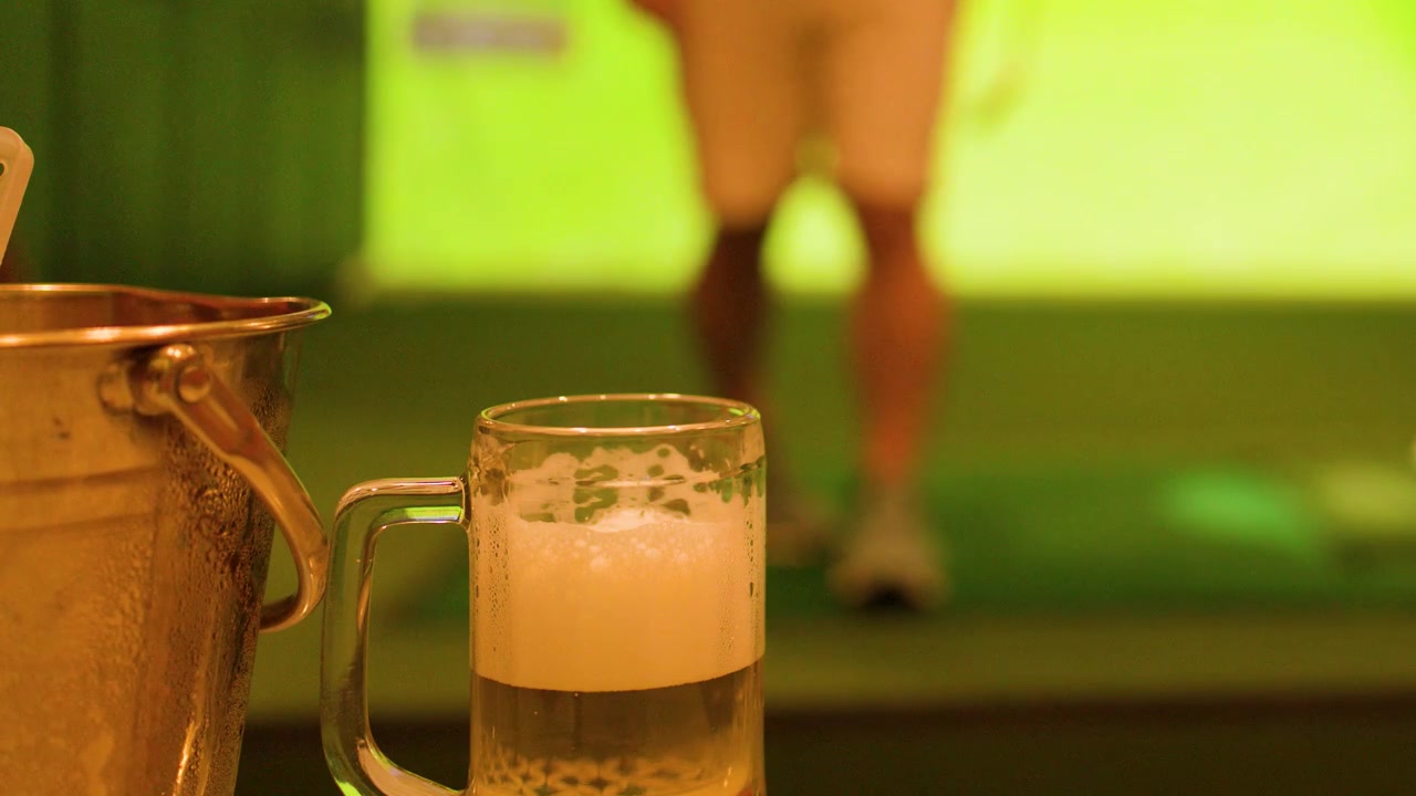 Statische Aufnahme von Bierkrug und Eiskübel, während ein Mann Golf in einem Indoor-Simulator übt