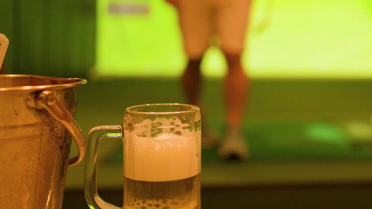 Inquadratura statica di un boccale di birra e un secchiello per il ghiaccio mentre un uomo si allena a golf in un simulatore indoor.