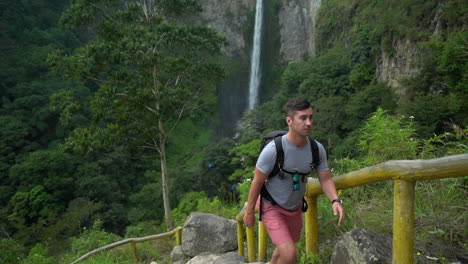 slow motion of a caucasian man hiking up a path with the flowing sipiso piso waterfall in the background in north sumatra