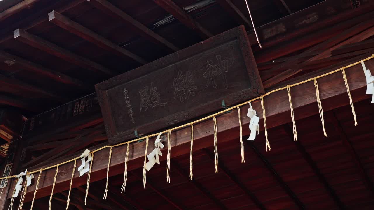 An old, weathered wooden signboard with intricate carvings hangs under the traditional Japanese roof. Shide (paper strips) adorn the ropes below, indicating a sacred space.