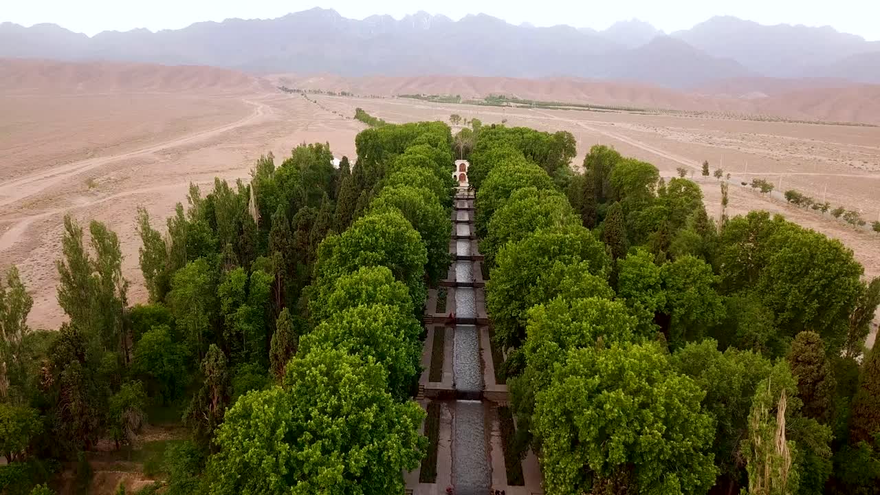 el tiro aéreo de un dron sube por el jardín fresco con árboles verdes, un arroyo y un hermoso palacio con un paisaje desértico y montañoso en irán