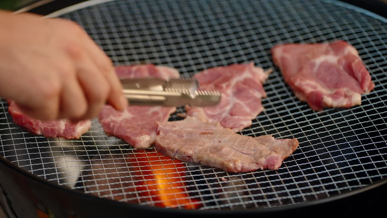 A close-up shot shows a hand using tongs to turn thin slices of pork as they sizzle on a wire mesh over hot charcoal, a classic method of cooking Korean barbecue (gogi-gui) at a camping site