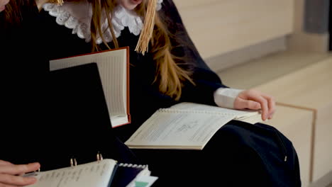 cerca de tres niñas felices con toga y birrete hablando y mirando sus cuadernos mientras se sientan en las escaleras en la ceremonia de graduación de preescolar