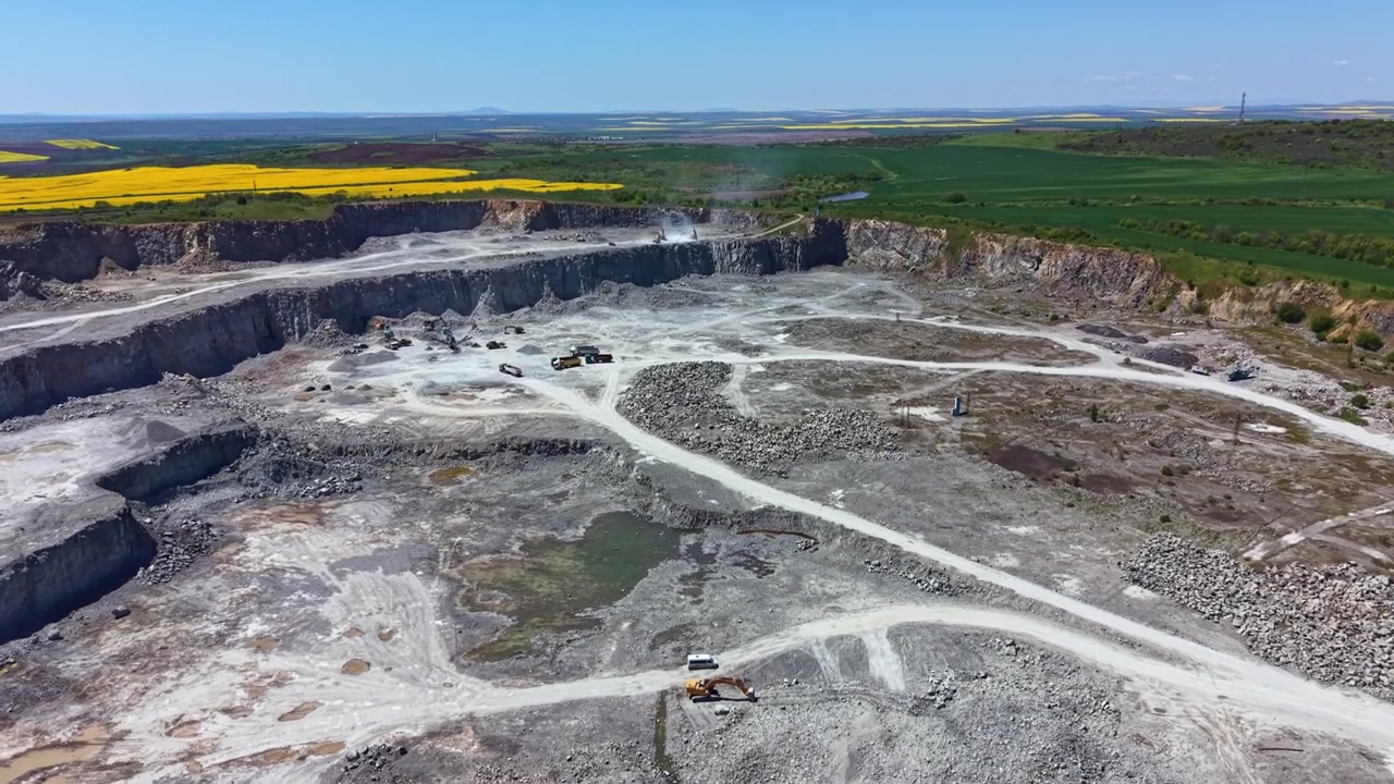 Macchinari pesanti lavorano in una grande cava in una zona rurale con campi verdi e cielo blu sullo sfondo durante le ore diurne.