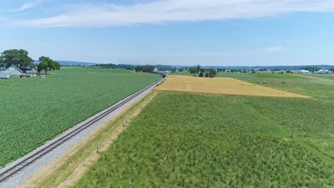 an aerial view of a steam engine puffing smoke and steam with passenger coaches traveling on a single track fertile farmland and countryside on a beautiful spring day