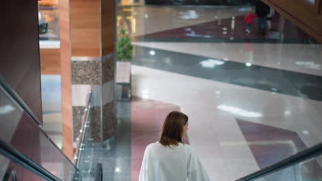 back view of young woman on moving escalator in brightly lit indoor shopping mall, showcasing polished floor patterns, modern architectural details with partial view of people in the down floor