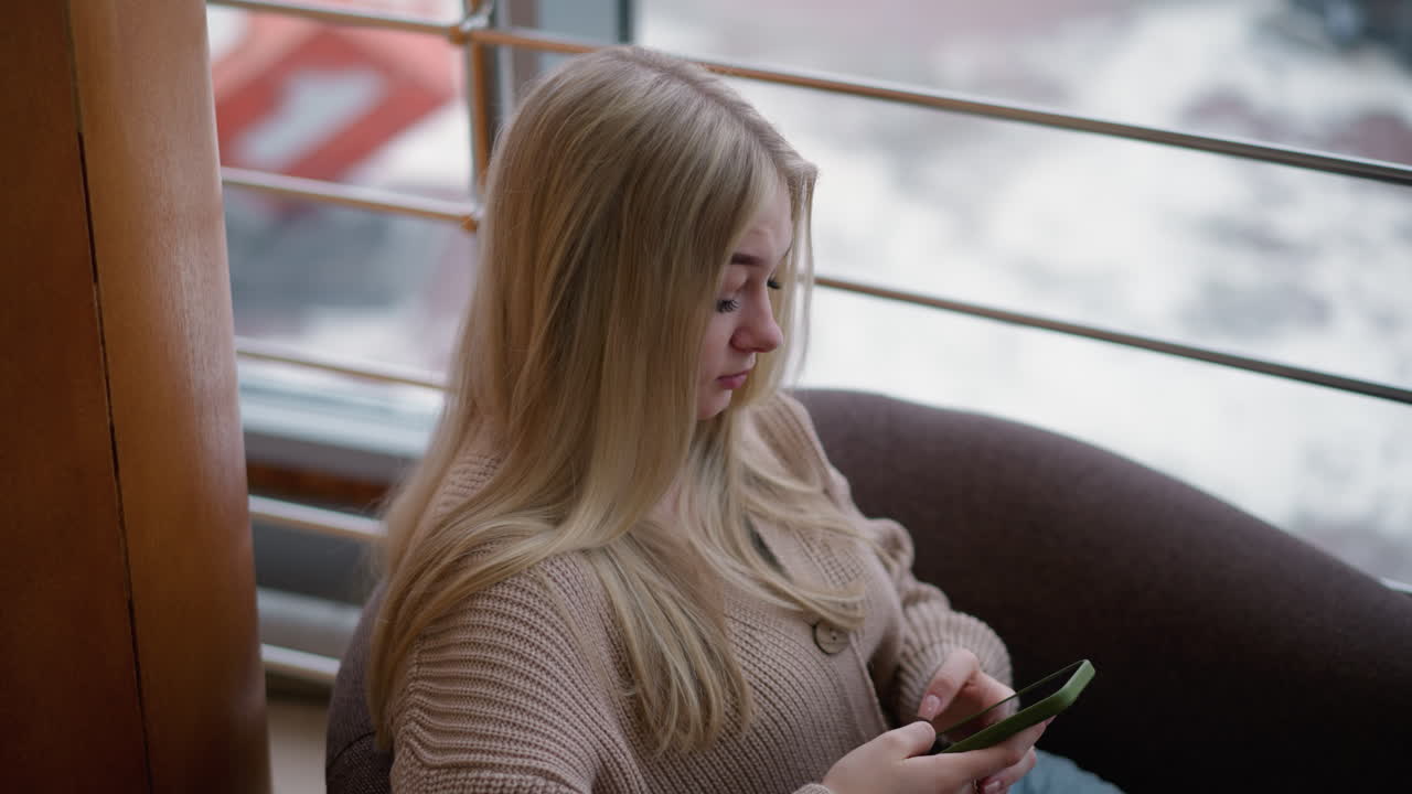 Nærbilde av en elegant kvinne med langt blondt hår, sittende i et moderne kjøpesenter, som ser tankefullt ut. Et urbant bybilde er synlig gjennom store vinduer med uskarpe snødekte gater.