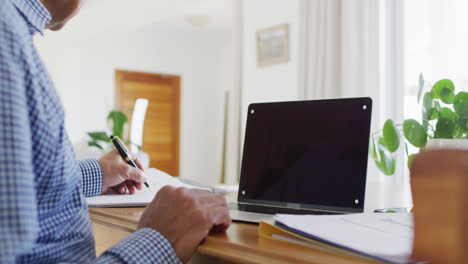 Video-of-senior-biracial-man-sitting-at-desk-writing-and-using-laptop-at-home,-copy-space-on-screen