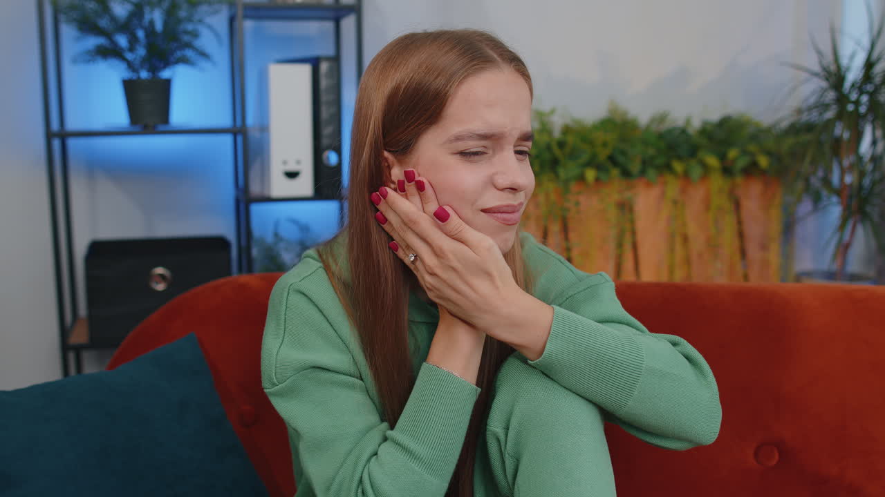 Ragazza che si tocca la guancia, sofferente per un acuto mal di denti, denti sensibili e carie, nella sua stanza di casa