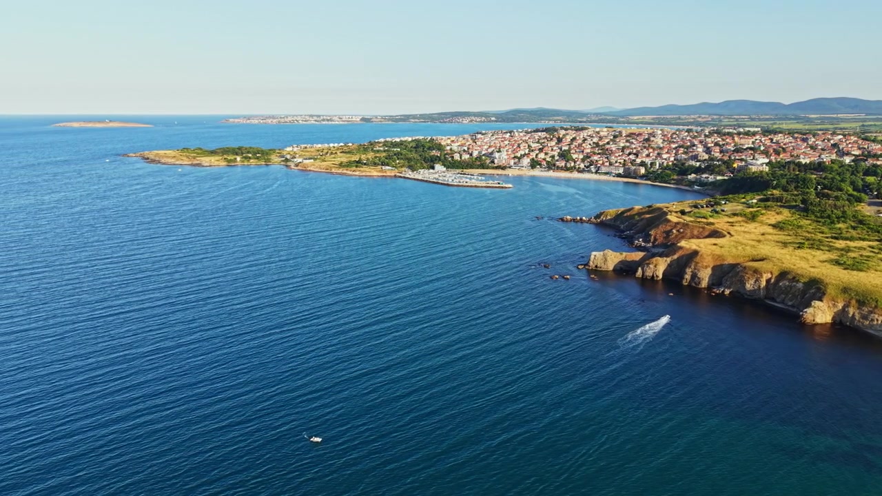 Vista costiera di un piccolo paese sul mare di giorno, con colline verdi e acqua in primo piano ed edifici sullo sfondo.