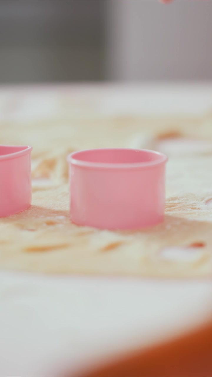 Close up view of pink plastic cookie cutters on rolled out dough with hands shaping background activity. scene focuses on baking preparation details like texture and material interaction