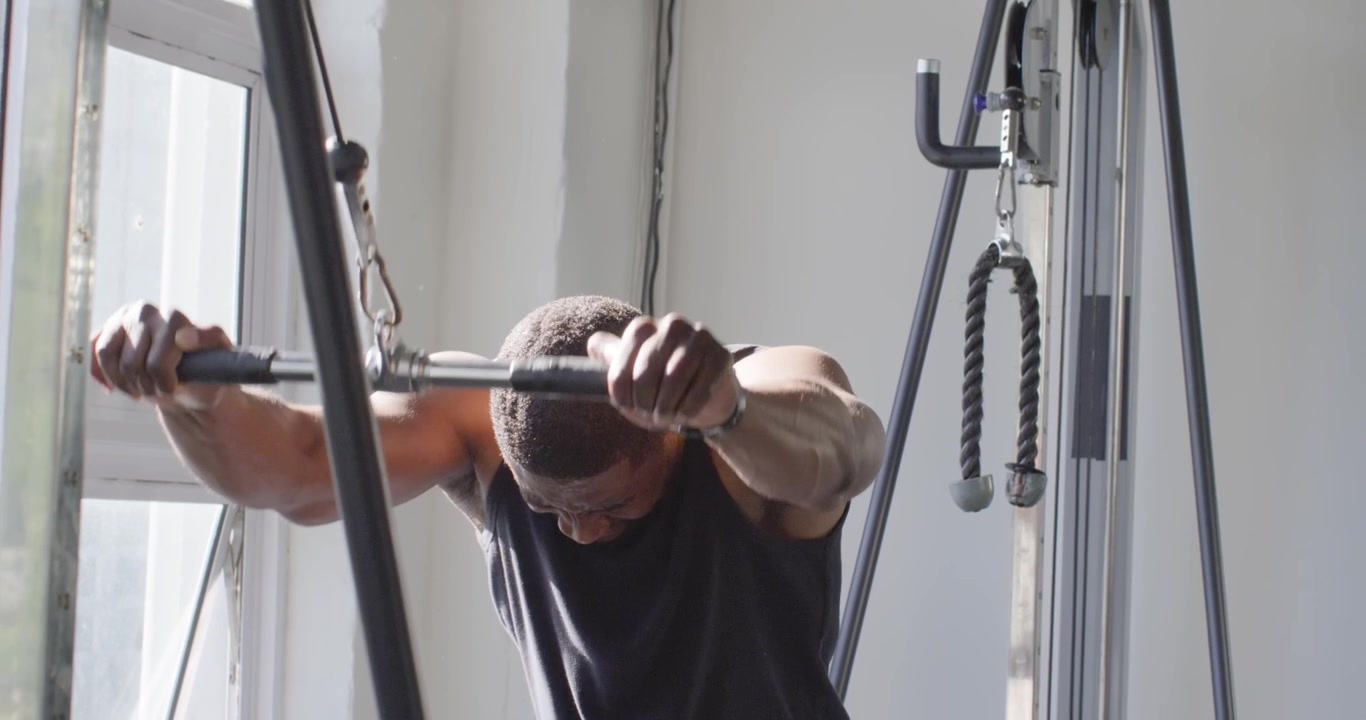 Uomo africano di mezza età che impugna una barra per cavi e tira verso il basso, sviluppando la forza della parte superiore del corpo in palestra.