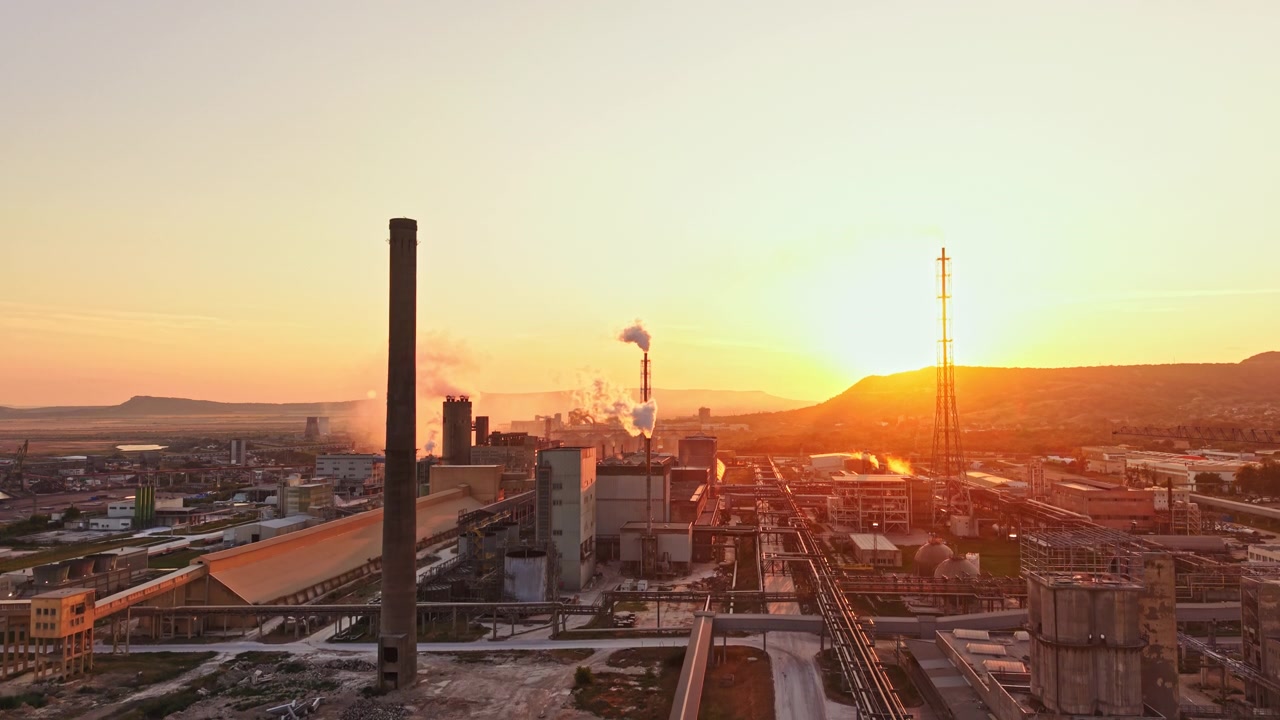 Indústria ao pôr do sol com fumaça e edifícios à vista perto de uma cordilheira.