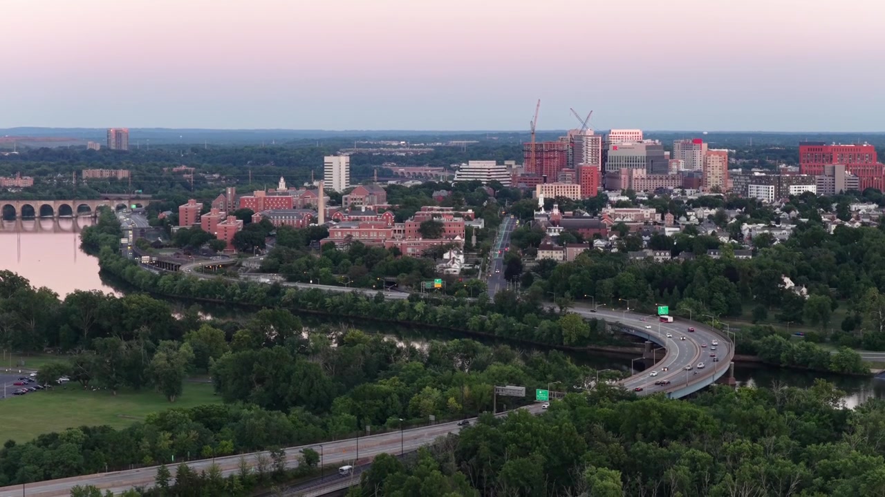 Virajlı otoyol, New Brunswick, New Jersey şehir merkezini geçerek Raritan Nehri'ni takip ediyor; arabalar yolda seyrederken kırmızı tuğlalı binalar ve şehir silüeti alacakaranlık ışığında yükseliyor. Havadan drone geniş çekim