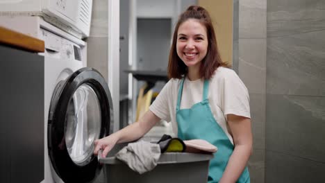 Retrato-De-Una-Señora-De-La-Limpieza-Morena-Segura-De-Sí-Misma-Con-Una-Camiseta-Blanca-Y-Un-Delantal-Azul-Que-Se-Sienta-Con-Un-Recipiente-De-Plástico-Lleno-De-Cosas-Sucias-Cerca-De-La-Lavadora-En-El-Baño-Mientras-Limpia-En-Un-Apartamento-Moderno.