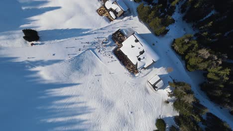 aerial: birds eye view, drone flying down to a ski-bungalow, surrounded by a slope with skking people