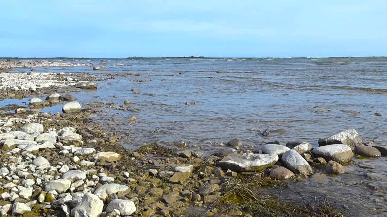 Gorgeous slow motion footage of a rocky beach shoreline during a sunny day where small Baltic ocean sea water waves and water ripples are coming to the shore and making rocks and pebbles wet, blue sky