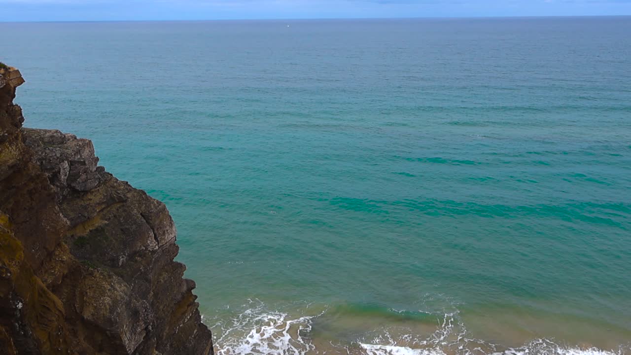 Vista diurna elevata da una drammatica scogliera portoghese che rivela onde atlantiche turchesi che si infrangono su una spiaggia sabbiosa e deserta sottostante.