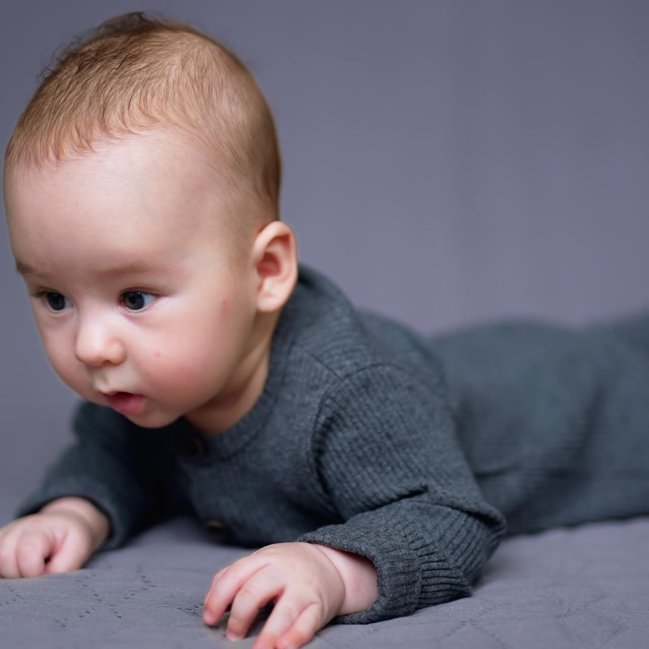Sweet Caucasian child in grey outfit lies on belly on the bed. Beautiful child looking around with surprise. Grey backdrop