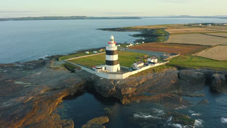 Vista-Aérea,-Salida-Del-Sol,-Tiro-Volador,-El-Faro-De-Gancho-Está-Situado-En-La-Cabeza-De-Gancho-En-La-Punta-De-La-Península-De-Gancho-En-Co-Wexford,-Irlanda,-El-Faro-Más-Antiguo-Del-Mundo,-Fue-Construido-En-El-Siglo-XII