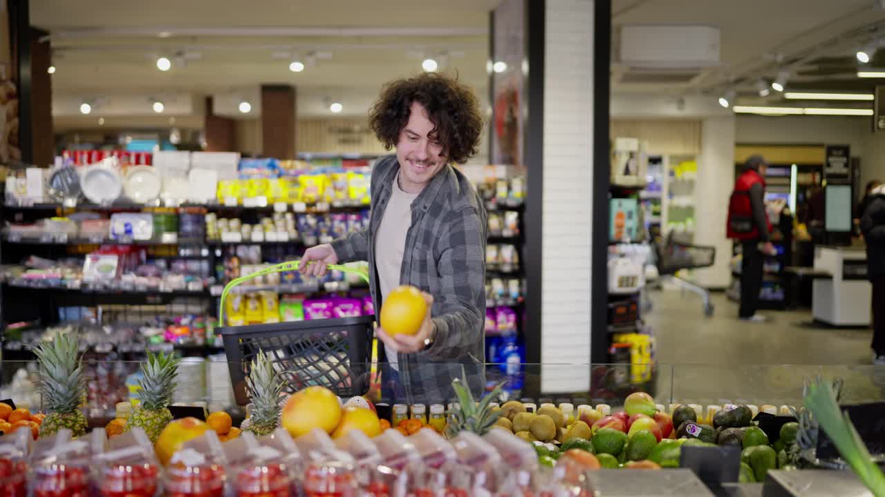 바구니를 가진 행복한 남자가 쇼핑하는 동안 슈퍼마켓에서 사탕수수 과일을 선택합니다. 식료품 가게에서 곱슬 머리카락을 가진 크레이드 셔츠를 입은 남자가