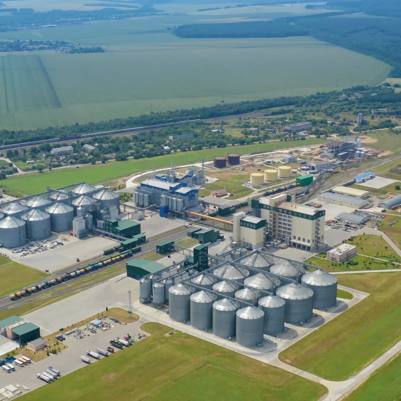 Modern granary surrounded by green nature. Storage tanks for keeping crop. Large agriculture complex on field. Silver grain elevators. Aerial view