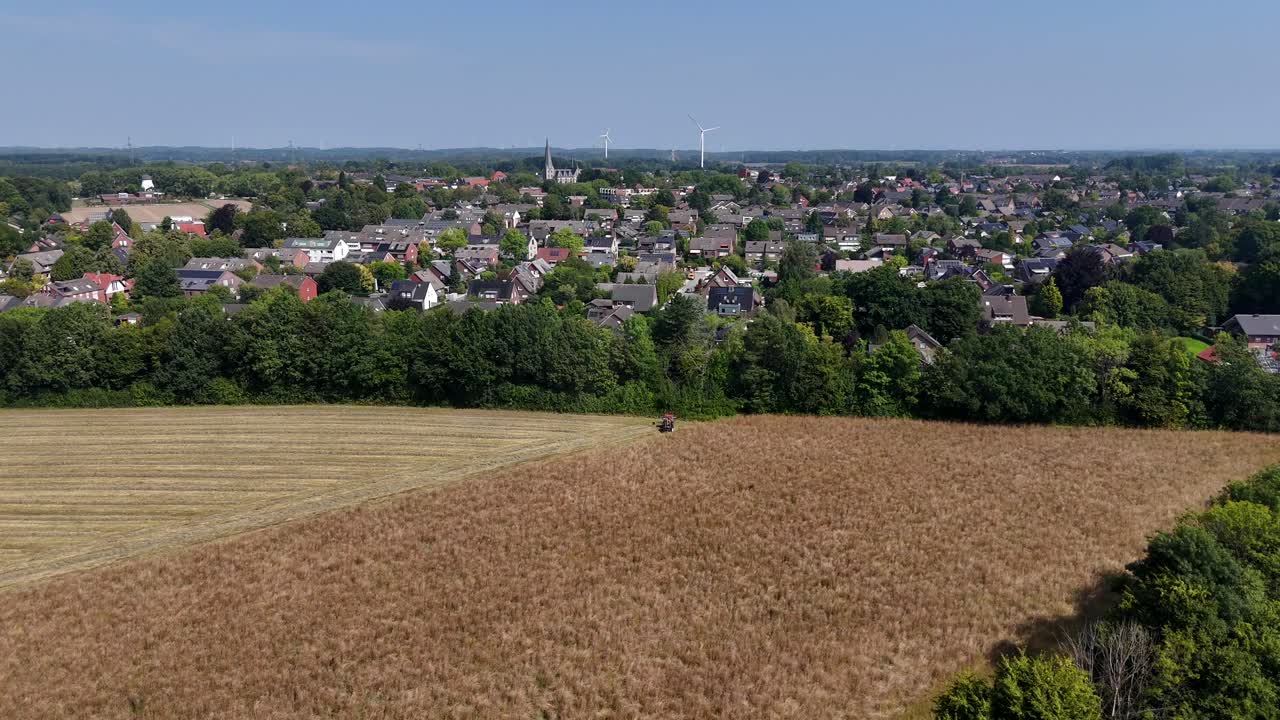 Tractor cutting on wheat field and harvesting corns on field. Aerial flyover shot. Cityscape and neighborhood with houses and wind turbines in background. Sunny day in summer