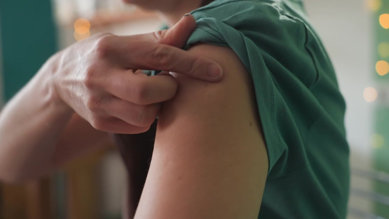 Closeup of arm rolling up sleeve, adult in green shirt prepares upper arm for injection in cozy room hand adjusts fabric, fingers touch skin, soft bokeh lights in background, calm anticipation