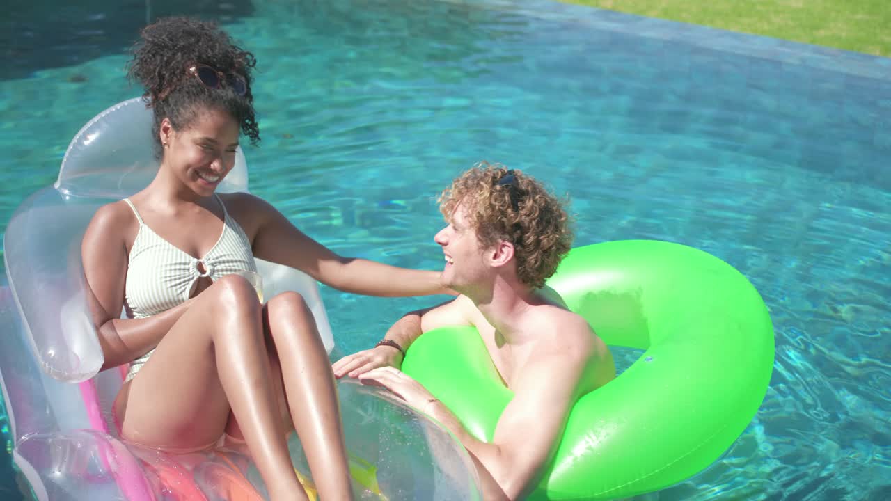 Diverse couple basking under sun on lounger and green tube at backyard pool, sharing citrus drink