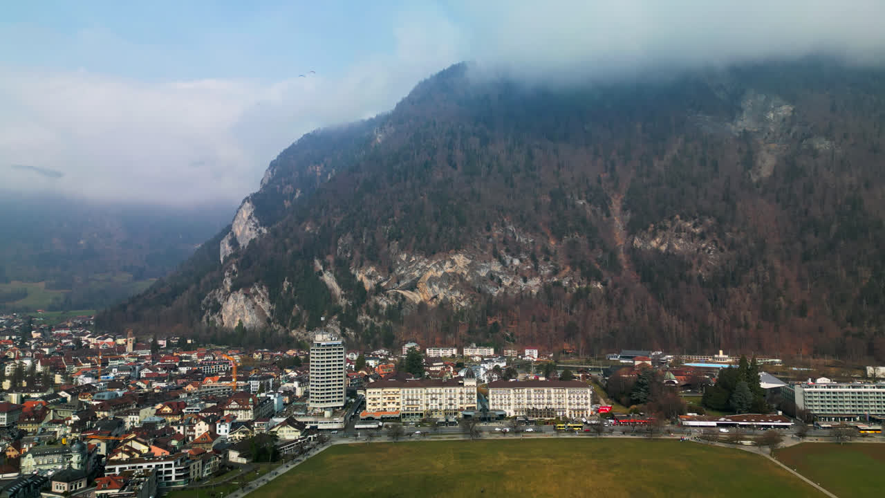 el dron asciende mostrando la extensa ciudad de interlaken, suiza, en la base de una montaña boscosa en las nubes.
