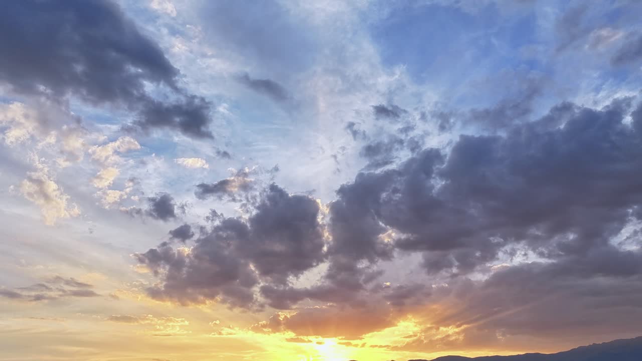 A drone hyperlapse at sunset shows the sun setting behind distant mountains. The sky is the star, with rapidly moving clouds and an intense light that gradually fades as day gives way to dusk