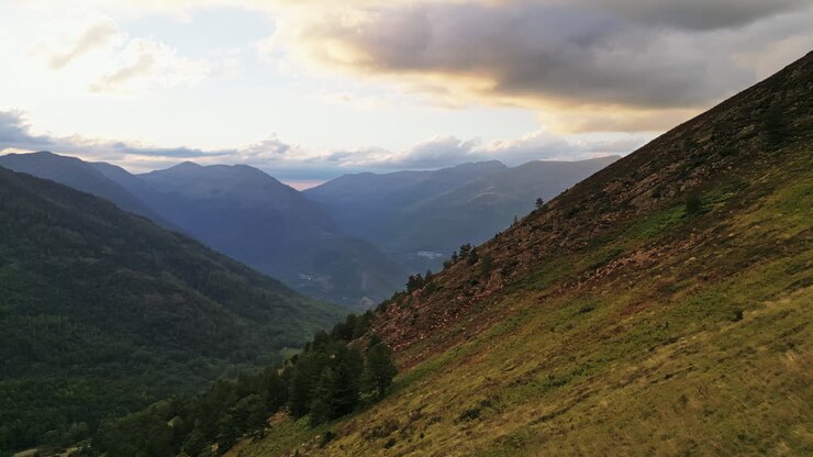 Toma de dron: Panorámica de un valle en las montañas con el atardecer al fondo, árboles en el suelo y nubes naranjas.