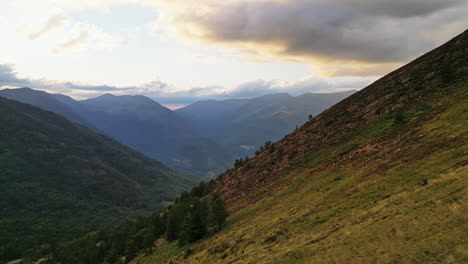 toma de drones: panorámica sobre un valle en las montañas con puesta de sol detrás, árboles en el suelo y nubes naranjas