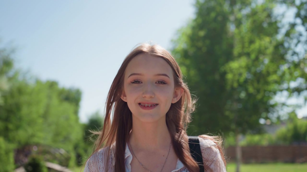 Primo piano di giovane donna che cammina all'aperto in una giornata di sole, con capelli lunghi e abiti leggeri, che porta uno zaino. I capelli si muovono naturalmente nel vento mentre la donna cammina lungo un sentiero circondato da alberi verdi.