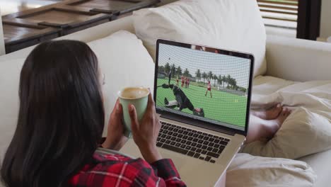 compuesto de una mujer sentada en casa sosteniendo un café viendo un partido de hockey en una computadora portátil