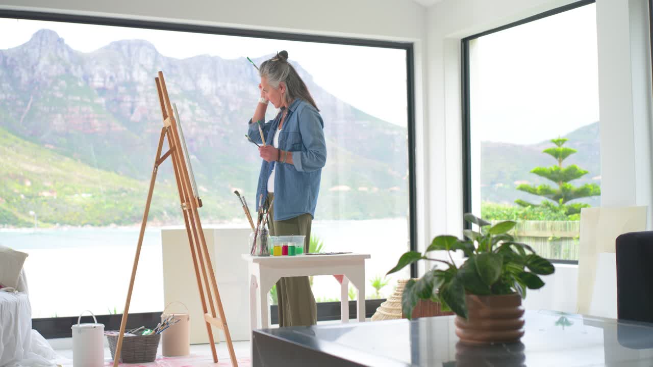 Eldre kvinne begynner med å dyppe pensler i malingskrukker, skaper kunstverk ved staffeliet ved vinduet.