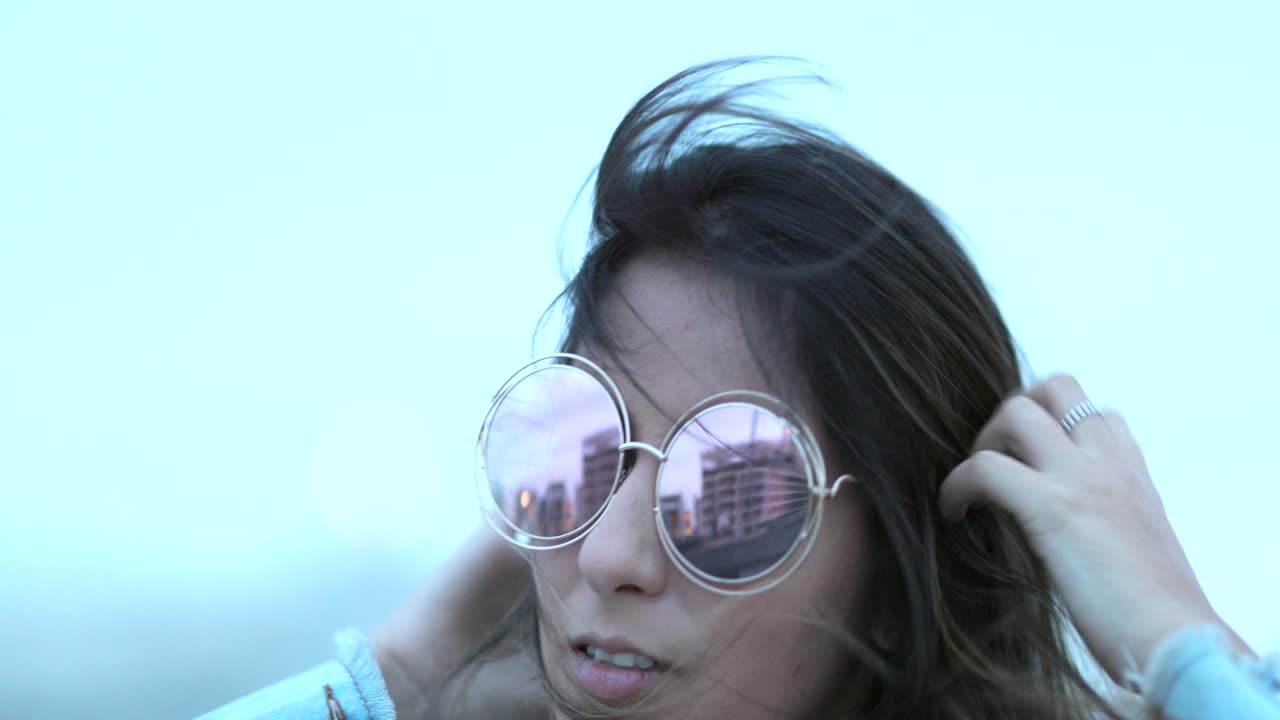 una mujer joven posando con grandes gafas de sol redondas y divertidas, el viento soplando el cabello