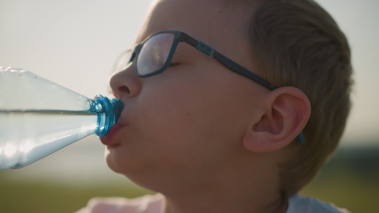 A close-up shot of a young boy in a white top, drinking water from a clear plastic bottle. His eyes are slightly closed, and he appears sleepy, enjoying a refreshing drink in a relaxed outdoor setting
