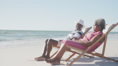 Happy-senior-african-american-couple-on-deck-chairs-and-talking-at-beach,-copy-space,-in-slow-motion